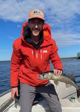 Mathieu portant un manteau rouge, debout sur un bateau et tenant un poisson